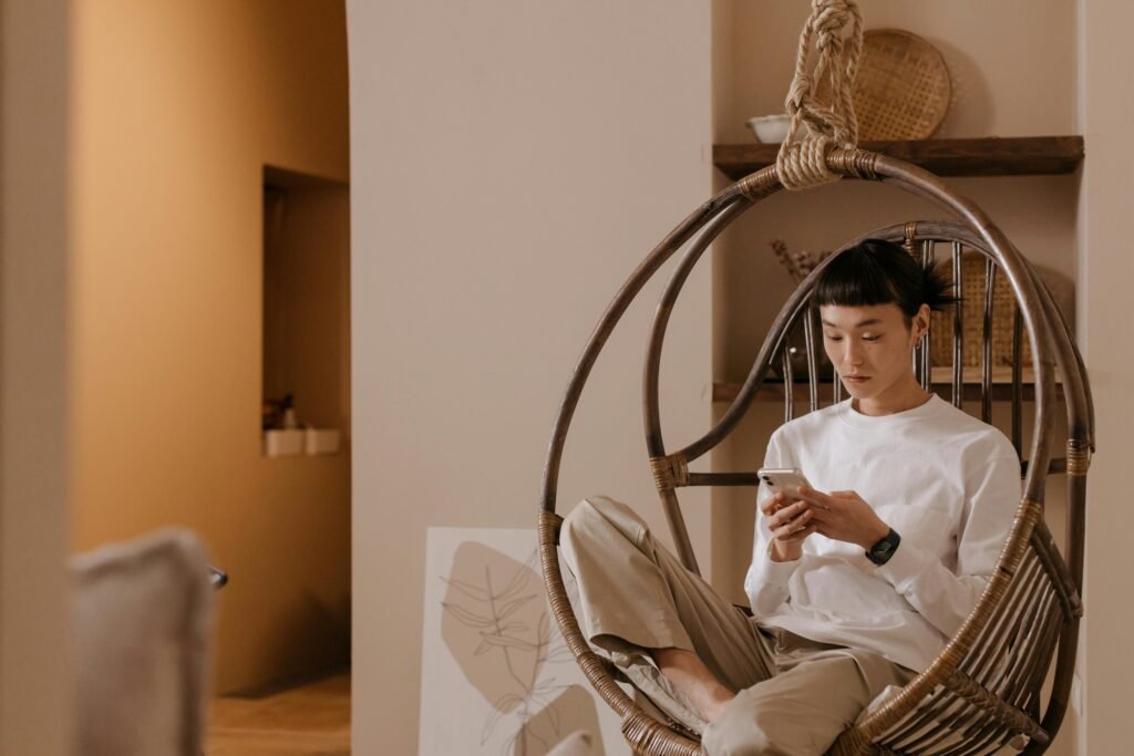 Asian man sitting in a hanging chair, texting on a smartphone indoors, wearing a white sweater.